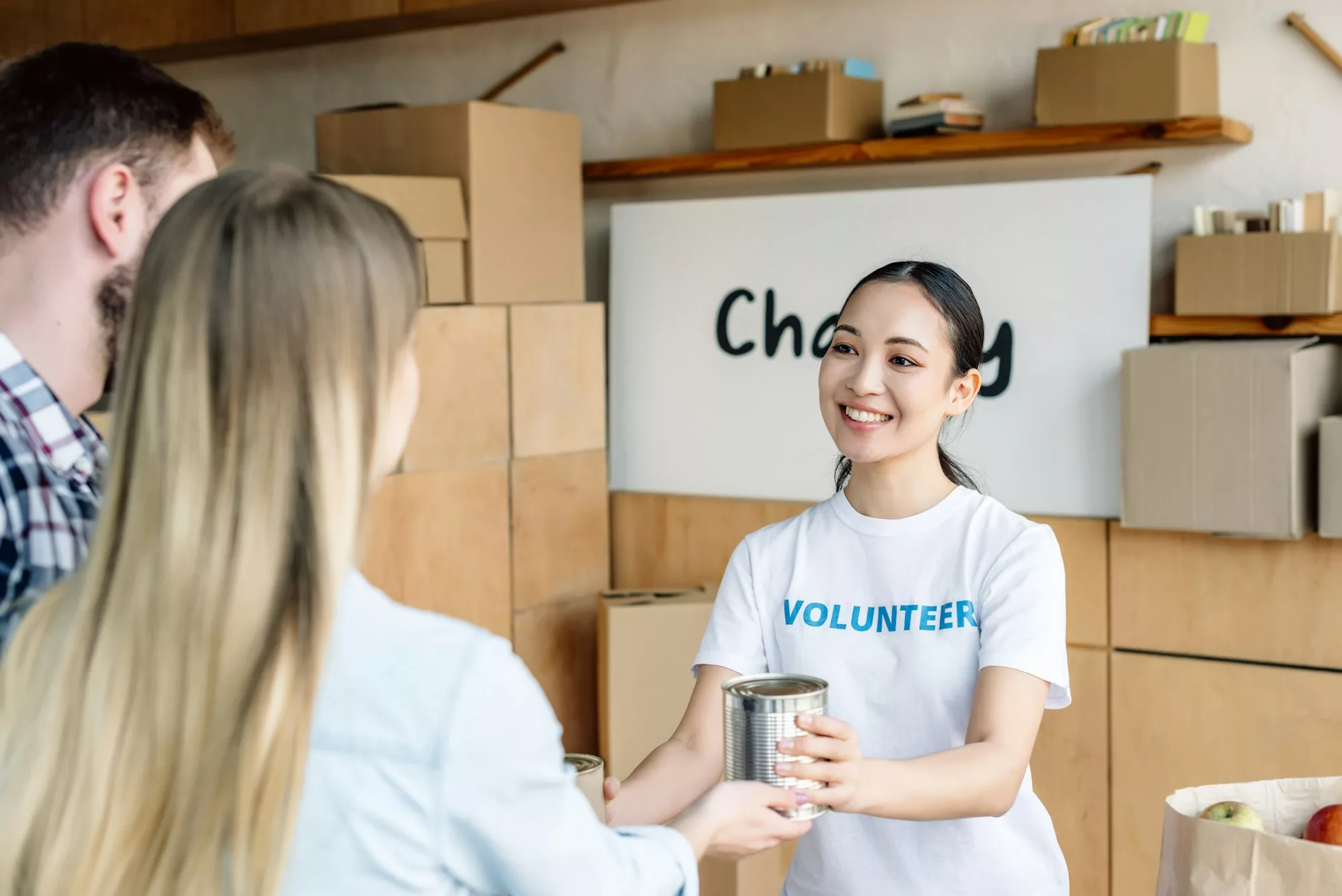 pretty asian volunteer giving canned food to man and woman in charity center